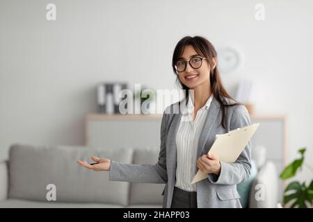 Glückliche Psychologin aus dem Nahen Osten mit Zwischenablage, die etwas mit der Hand zeigt, die Kamera anlächelt, den Raum kopiert Stockfoto