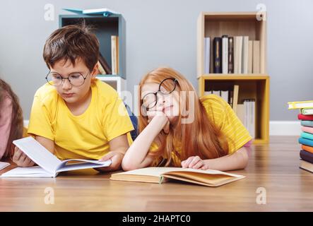 Niedliche Kinder in Gläsern, die drinnen mit Büchern auf dem Boden liegen Stockfoto