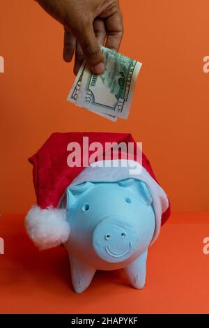 Hand setzen Dollar Geld in Sparschwein mit weihnachtsmann Hut isoliert auf orangefarbenem Hintergrund. Stockfoto