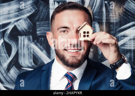 Nahaufnahme eines Immobilienmaklers oder Versicherers in Jacke und Krawatte. Der 25-35-jährige Kaukasusjunge hält ein Spielzeughaus in der Hand und lächelt. Hintergrund. Stockfoto