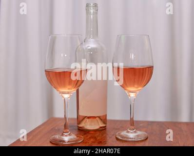 Flasche Roséwein mit unbeschrifttem Etikett mit zwei Gläsern auf Holztisch mit weißem Vorhang im Hintergrund. Vertikal. Verkostung von Roséwein. Paar, sehr junge Leute Stockfoto