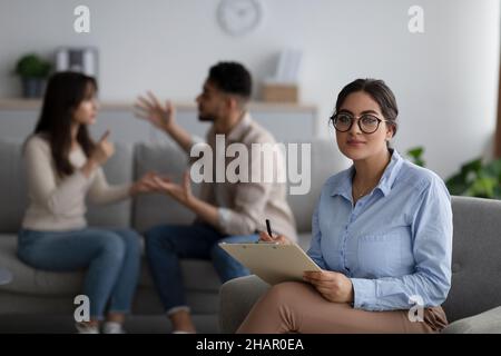 Heiratstherapie. Seriöser Familienberater, der die Kamera anschaut, während junge Ehepartner emotional argumentieren, selektiver Fokus Stockfoto