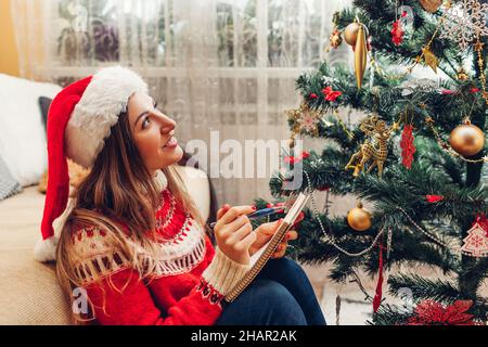 Junge Frau, die Vorsätze für 2022 schreibt, sitzt zu Hause im Notizbuch am Weihnachtsbaum und trägt einen Weihnachtsmütze und einen warmen roten Pullover. Motivation für Stockfoto
