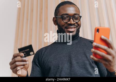 Fröhlicher afroamerikanischer Geschäftsmann mit Kreditkarte und Smartphone zum Einkaufen im Internet Stockfoto