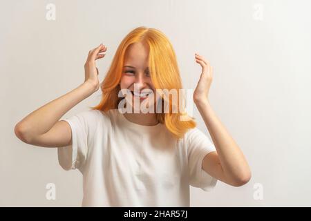 Jugendliches Mädchen mit roten Haaren in einem weißen T-Shirt auf hellem Hintergrund lacht und richtet das Haar auf. Stockfoto
