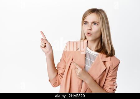 Verwirrt kleines Kind, blonde niedliche Mädchen zeigt und schaut links mit staunenden Gesicht, stehend in formalen Blazer vor weißem Hintergrund Stockfoto