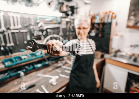 Porträt einer schönen kaukasischen Arbeiterin, die einen Schraubenschlüssel hält, während sie in einer Fahrradwerkstatt steht. Selektiver Fokus auf Schraubenschlüssel. Stockfoto