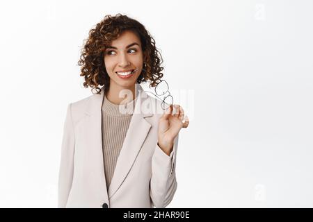 Erfolgreiche, attraktive Geschäftsfrau, die mit einer Brille steht, eine Brille hält und glücklich lächelt und auf weißem Hintergrund in beigefarbenem Anzug steht Stockfoto