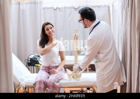 Weibliche Patientin im Schlafanzug sitzt auf dem Krankenhausbett und zeigt einen schmerzenden Platz am Hals. Doktor, der neben ihr steht und ein Wirbelsäulenmodell hält. Stockfoto