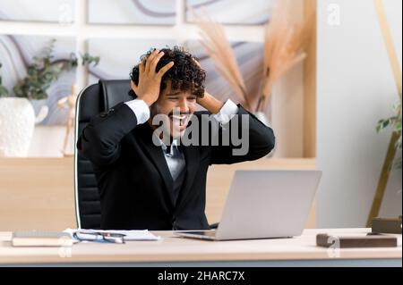 Frustriert junger, erwachsener ceo, Geschäftsmann oder Manager aus Indien, mit einem Bart, sitzt an einem Tisch im Büro, frustriert über finanzielle Verluste, Zusammenbruch, gestresst mit Handgesten, schreit Stockfoto