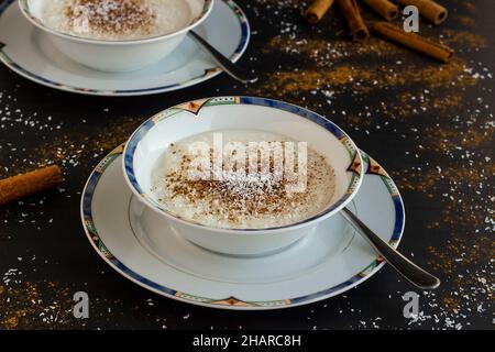 Traditioneller türkischer Reispudding Sutlac im edlen Dessert-Set aus Porzellan, auf der schwarzen Oberfläche mit Zimtstangen und Kokosnusspulver. Stockfoto