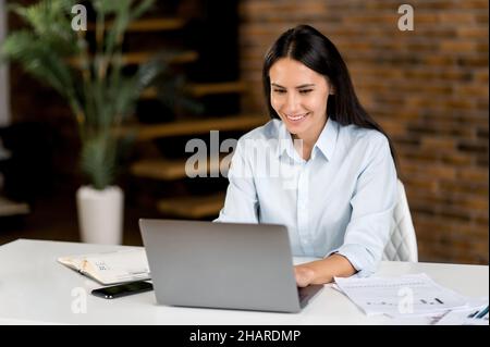 Niedlich erfolgreiche smart freundlich kaukasisch brünette, top-Manager, Broker, sekretärin, real estate Agent, Arbeiten mit einem Laptop, Sitzen am Tisch im Büro, SMS mit Kunden oder Kollegen, lächelnd Stockfoto