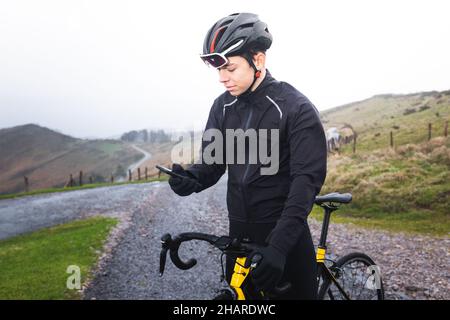 Junger kaukasischer Radfahrer, der ein Smartphone auf seinem Fahrrad auf dem Gipfel eines Berges benutzt. Stockfoto