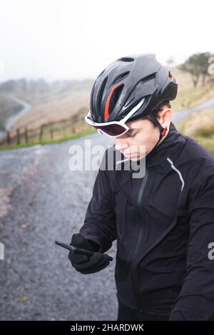 Junger kaukasischer Radfahrer, der ein Smartphone auf seinem Fahrrad auf dem Gipfel eines Berges benutzt. Stockfoto