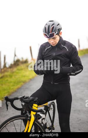 Junger kaukasischer Radfahrer, der ein Smartphone auf seinem Fahrrad auf dem Gipfel eines Berges benutzt. Stockfoto