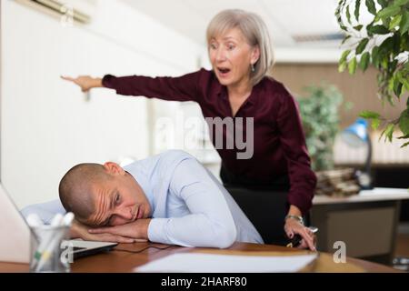 Büroangestellte schläft am Arbeitsplatz vor dem Hintergrund einer unzufriedenen Chefin Stockfoto