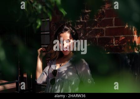 Ein Porträt zwischen den Blättern eines Busches einer schönen jungen Frau, die sich an einer Wand lehnt. Stockfoto