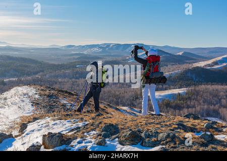 Ein Paar mittleren Alters fotografiert mit dem Smartphone, während es bei Sonnenaufgang in den alpinen Winterbergen wandert. Stockfoto