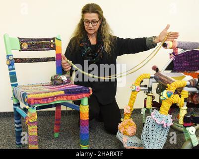 14. Dezember 2021, Sachsen, Torgau: Sozialarbeiterin Ina Baumann-Kahlo hält in ihrem Büro neben einem Kinderrollstuhl, der mit farbiger Wolle (r) eingestrickt ist, einen „Patience Thread“ und einen Stuhl, der ebenfalls in ihren Händen eingestrickt ist. Während der Corona-Pandemie habe Baumann-Kahlo viele Stunden im Rollstuhl gestrickt, um den vielen Helfern in der Krise zu danken, die in der Öffentlichkeit nicht so präsent waren, sagte sie. Die Menschen sehen den Rollstuhl, wie sie es tut, als ein Symbol für das Bedürfnis nach Hilfe. Sie sieht den Stuhl als einen 'Helferstuhl'. Damit möchte sie sich bei den vielen Helfern bedanken, die anderen still und leise helfen, auch in Stockfoto