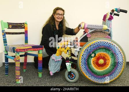 14. Dezember 2021, Sachsen, Torgau: In ihrem Büro zeigt die Sozialarbeiterin Ina Baumann-Kahlo einen Kinderrollstuhl, der mit farbiger Wolle (r) eingestrickt ist, und einen Stuhl, der ebenfalls eingestrickt ist. Während der Corona-Pandemie hat Baumann-Kahlo viele Stunden im Rollstuhl gestrickt, als Dankeschön an die vielen Helfer in der Krise, die in der Öffentlichkeit nicht so präsent sind. Die Menschen sehen den Rollstuhl, wie sie es tut, als ein Symbol für das Bedürfnis nach Hilfe. Sie sieht den Stuhl als einen 'Helferstuhl'. Damit möchte sie sich bei den vielen Helfern bedanken, die auch in der Corona-Krise still anderen helfen. Foto: Waltraud Grubitzsch/dpa-Zentralbi Stockfoto