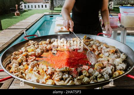 Prozess der Zubereitung von Paella in einer Pfanne mit Huhn und Gemüse und Köchin's Hand würzen und Kochen auf einer Terrasse in Spanien Stockfoto