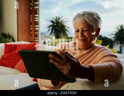 Glückliche multikulturelle ältere Frau, die ein elektronisches Tablet benutzt und tagsüber auf dem Sofa im Wohnzimmer sitzt. Stockfoto