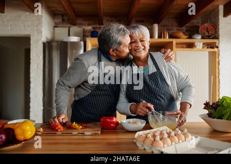 Biracial ältere Paar glücklich Kochen in der Küche. Ehemann küsst Frau auf die Wange, gesunder Lebensstil im Ruhestand. Stockfoto