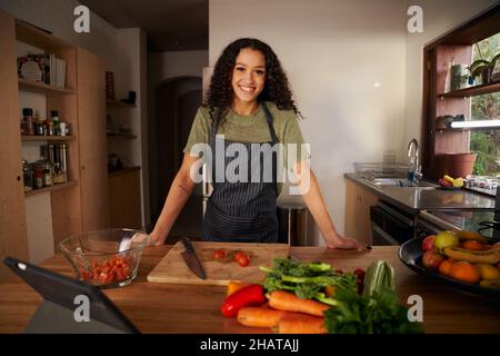 Junge, schöne afroamerikanische Frau, die lächelt, während sie in einem Kätzchen mit Schürze beim Kochen steht Stockfoto