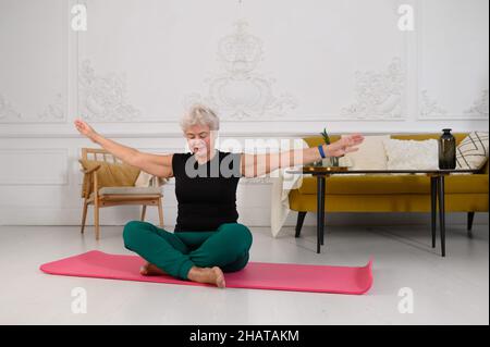 Eine schön aussehende, gealterte Frau macht zu Hause Yoga in einem wunderschönen weißen Interieur Stockfoto