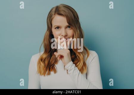 Lächelnde junge Millennials-Frau, die ihren Zeigefinger auf den Lippen über dem blauen Hintergrund der Studiowand hält Stockfoto