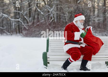 Älterer Mann mit rotem Weihnachtsmann-Kostüm sitzt am verschneiten Wintertag auf der Bank im Park und bereitet sich darauf vor, Geschenke in den Sack zu stecken Stockfoto