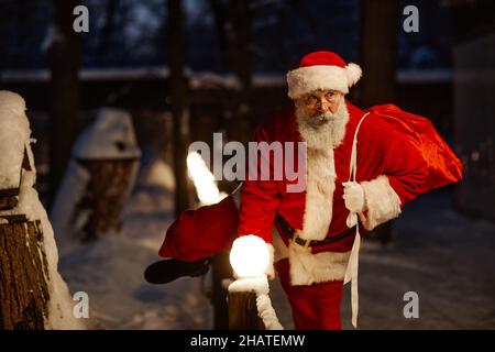 Horizontale mittellange Porträtaufnahme des modernen Weihnachtsmanns, der leise über den Zaun tritt, um nachts Geschenke zu liefern Stockfoto