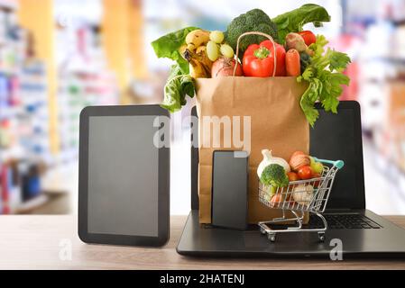 Online-Shopping-Konzept mit mobilen Geräten und Supermarkt-Einkaufstasche voller Obst und Gemüse auf dem Tisch im Supermarkt. Vorderansicht. Horizontal Stockfoto