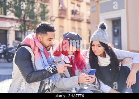 Stilvolle Frau mit langen roten Haaren, die ein Smartphone mit multirassischen Freunden teilt und auf der Straße in der Stadt gemeinsam auf den Bildschirm schaut Stockfoto