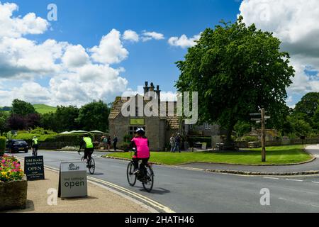 3 Radfahrer und Spaziergänger, die an einem malerischen, attraktiven Cottage Tea Rooms Cafe in einem malerischen, sonnigen ländlichen Dorf vorbeikommen - B6160 Bolton Abbey, Yorkshire Dales, England. Stockfoto