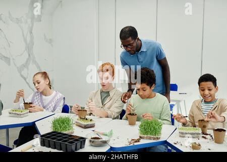 Verschiedene Gruppe von Kindern, die Samen Pflanzen, während sie in der Biologie-Klasse in der Schule mit afroamerikanischem Lehrer experimentieren, der beaufsichtigt Stockfoto
