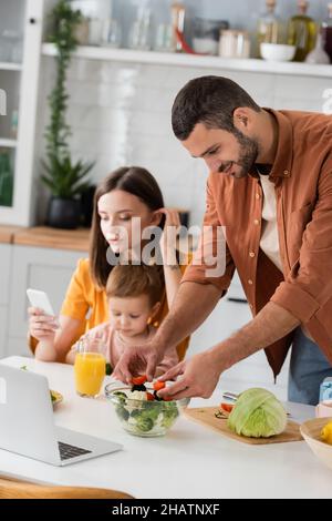 Mann kocht Salat in der Nähe des Laptops und verschwommene Familie in der Küche Stockfoto