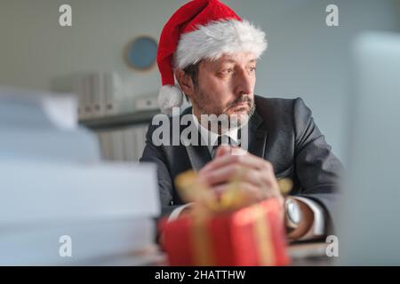 Besorgter Geschäftsmann mit Weihnachtsmann-Hut, der während der Weihnachtsfeiertage aus dem Bürofenster schaut, selektiver Fokus Stockfoto