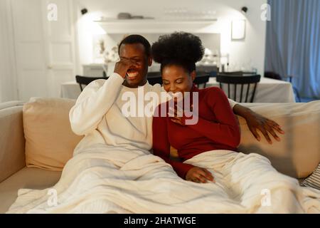 Glückliches junges afroamerikanisches Paar, das über einen lustigen Witz lacht und sich gemeinsam zu Hause einen Comedy-Film ansieht Stockfoto
