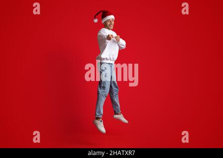 Fröhlicher Mann Mit Weihnachtsmütze, Der In Der Luft Springt Und Auf Die Kamera Zeigt Stockfoto