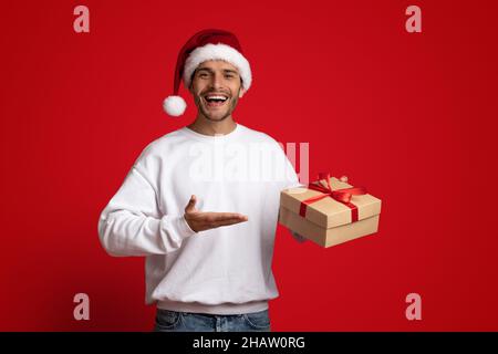 Fröhlicher Mann Mit Weihnachtsmütze, Der In Seiner Hand Auf Die Geschenkbox Zeigt Stockfoto