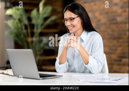 Freundliche, hübsche, erfolgreiche kaukasische Brünette Geschäftsfrau, Maklerin, Unternehmensmanagerin, die einen Laptop benutzt, während sie am Schreibtisch sitzt, ein Projekt plant, gestikelt und glücklich lächelt Stockfoto