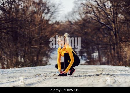 Die Sportlerin hockt im Winter auf verschneiten Pfaden in der Natur und schnürt ihr Schuhwerk. Sportbekleidung, Winterfitness, gesundes Leben Stockfoto