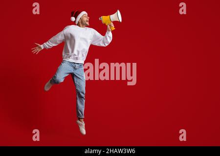 Ankündigung Zu Weihnachten. Aufgeregt Junger Mann In Santa Hat Schreiend Mit Lautsprecher Stockfoto