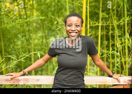 Attraktive glücklich lächelnde junge natürliche Schönheit kurzhaarige afrikanische Schwarze Frau lachend lehnt sich an ein Holzgeländer posiert vor dem Hintergrund der Bambushain im Natur Sommerpark Stockfoto