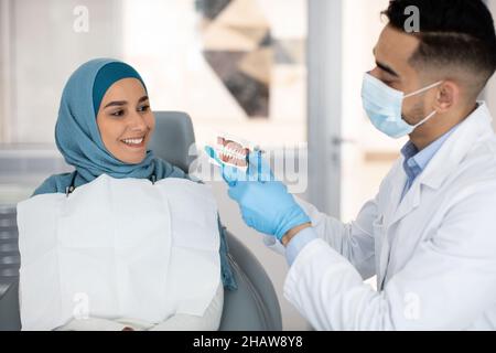 Männlicher Zahnarzt mit Kiefermodell, erklärt der muslimischen Patientin die Zahnhygiene Stockfoto