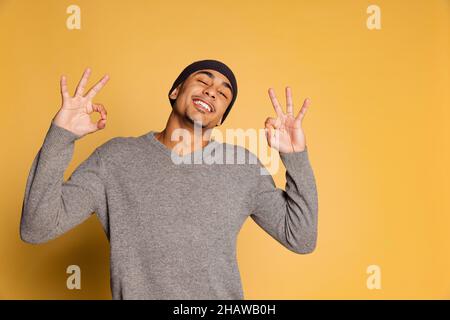 Halblanges Porträt eines afrikanischen jungen Mannes in warmen Kleidern und Hut isoliert auf gelbem Studiohintergrund. Konzept der menschlichen Emotionen Stockfoto