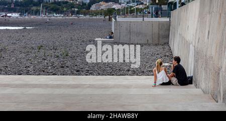 Junges Paar, das auf einer Betontreppe an einem Kiesstrand in Funchal, Madeira, Portugal sitzt Stockfoto