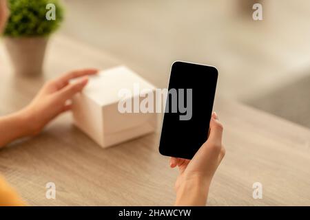 Frau hält Handy mit leerem Bildschirm für Mockup, schreiben Bewertung für Produkt, berühren Karton Stockfoto