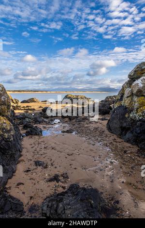 Ein felsiger Strand bei Ebbe auf Ynys Llanddwyn mit den Snowdonia-Bergen über der Bucht, Isle of Anglesey, Nordwales Stockfoto